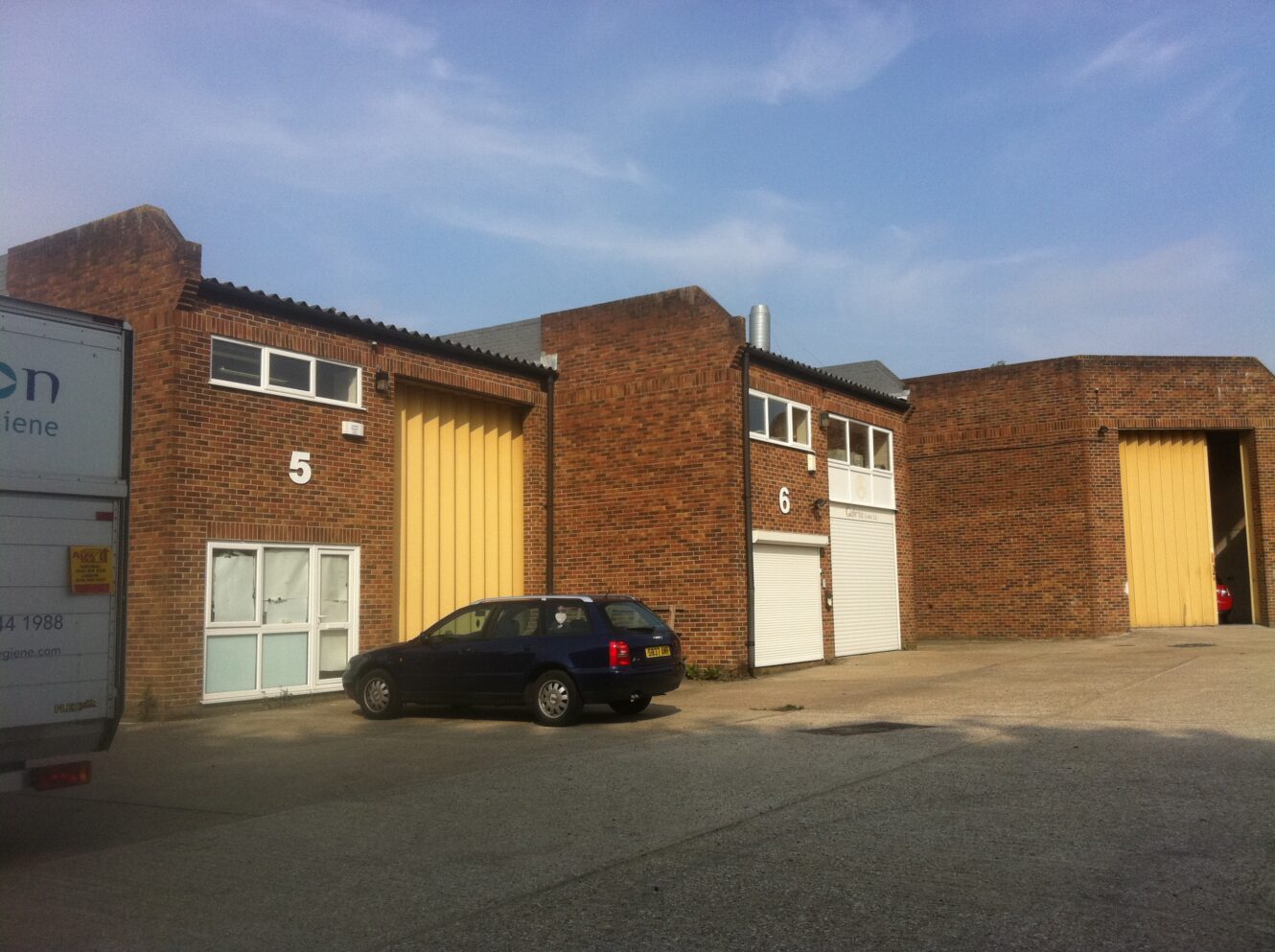 Brick industrial units numbered 5 and 6 with yellow doors, a black car parked in front, and part of a truck on the left under a clear sky.
