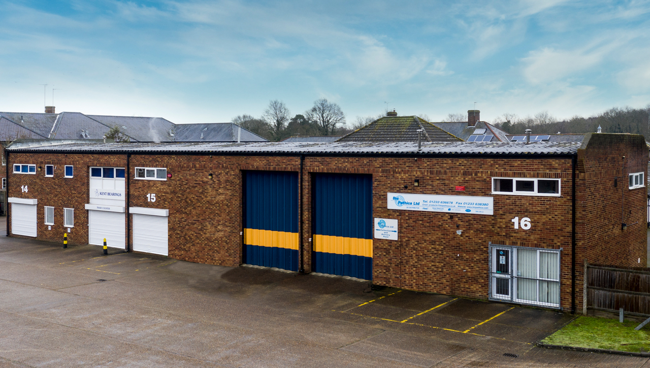 A row of brick industrial warehouse units with large roller doors, numbered 14, 15, and 16, with clear parking spaces in front under a partly cloudy sky.