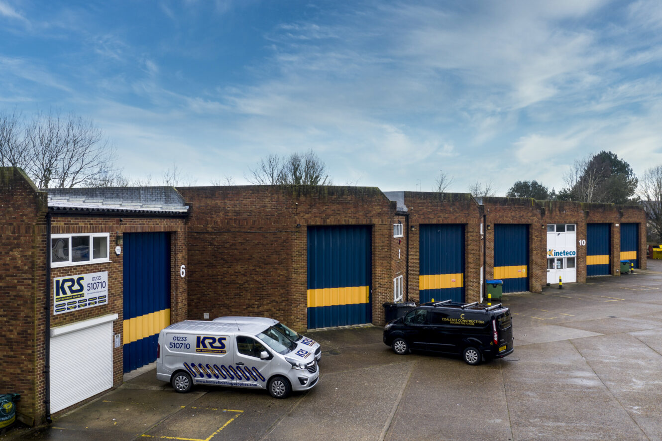 A row of brick industrial warehouse units with blue and yellow doors, two branded vans parked in front, and a partly cloudy sky overhead.
