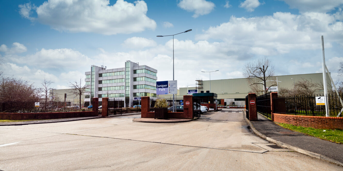 Entrance to a commercial or industrial park with office and warehouse buildings, parking spaces, and signage for “Juniper Park” under a partly cloudy sky.