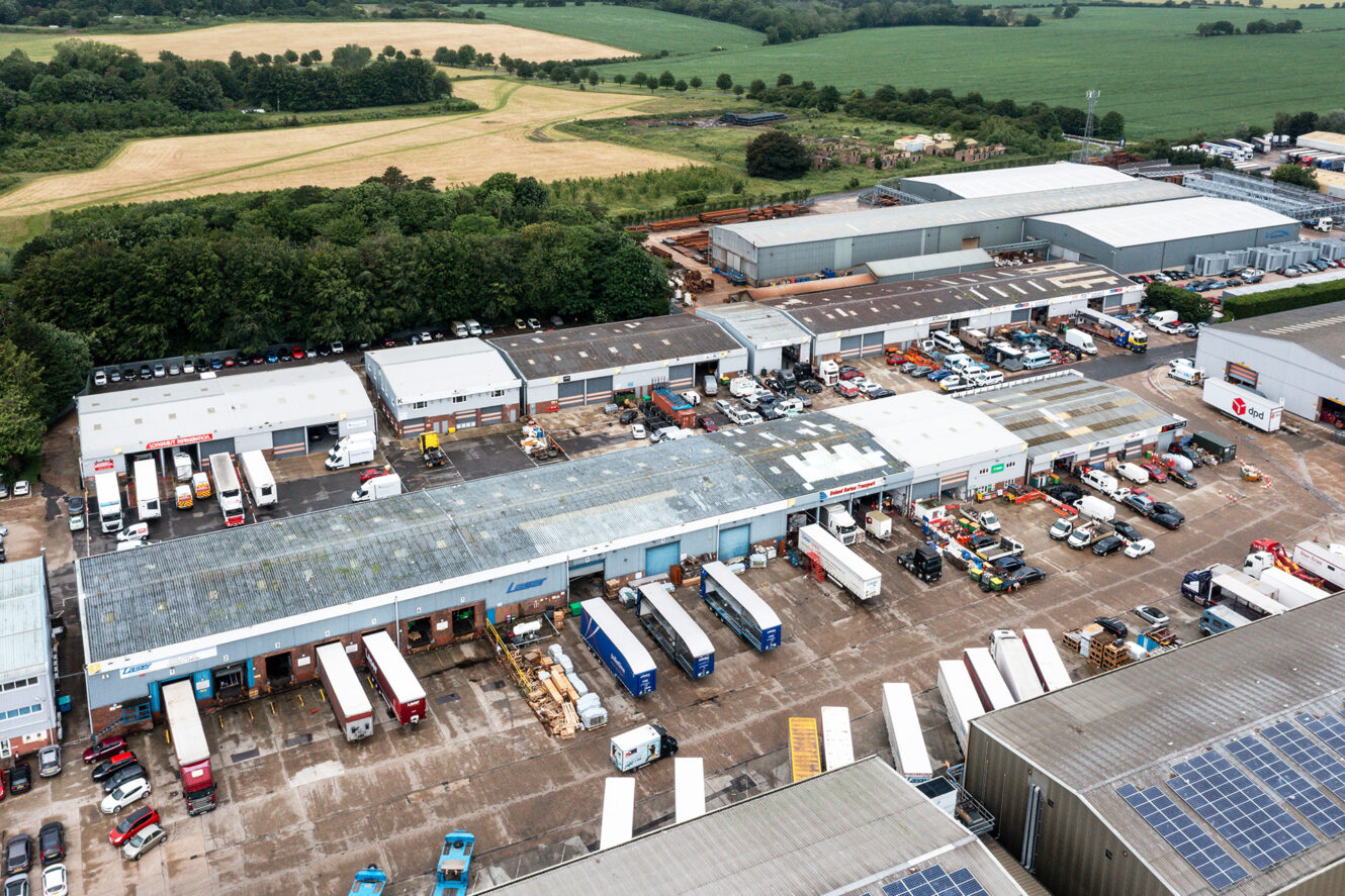 Aerial view of an industrial park with multiple warehouses, trucks, parked vehicles, and surrounding green fields.