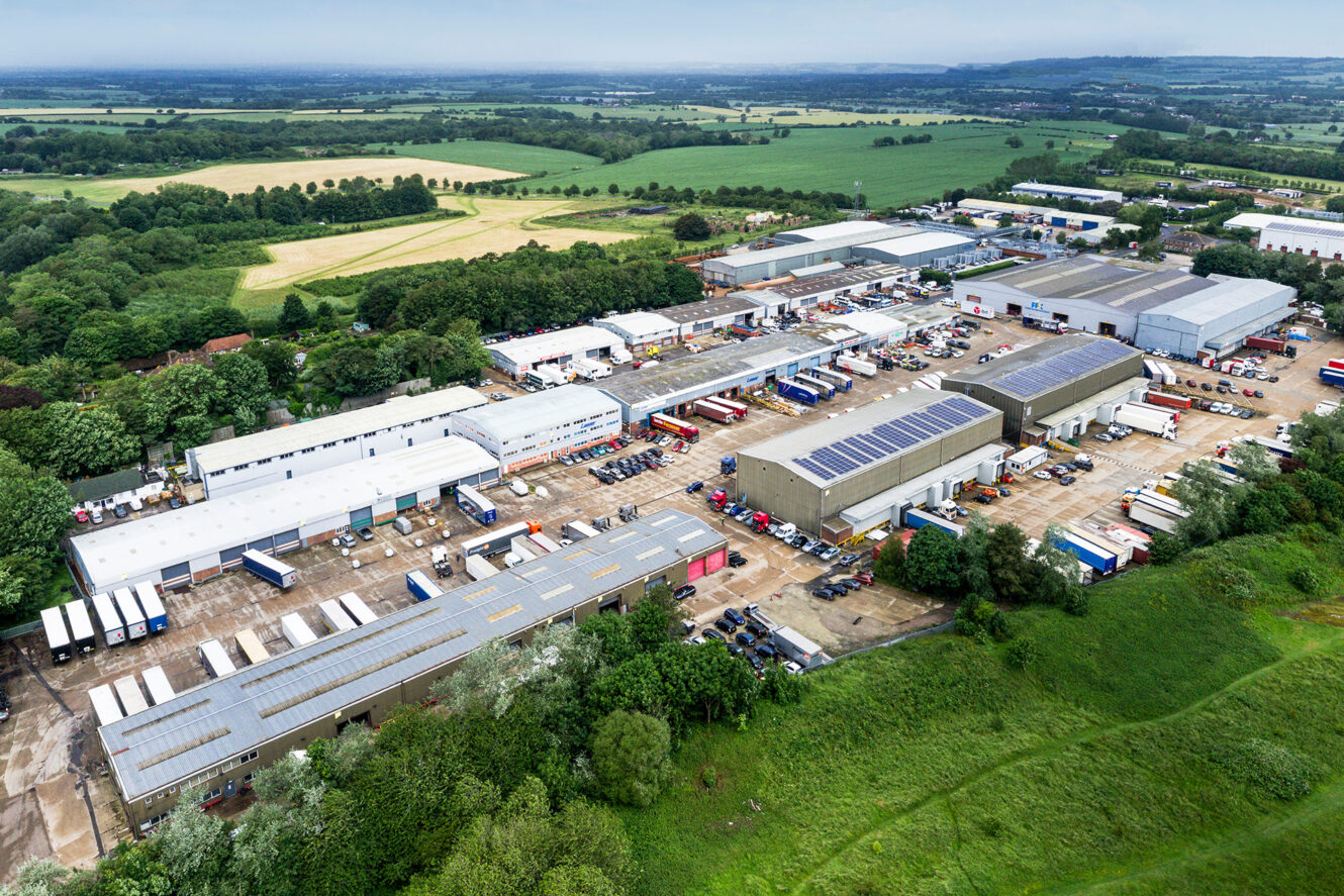 Aerial view of an industrial estate with warehouses, parked trucks, and surrounding green fields on a cloudy day.