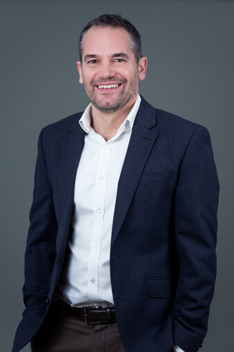 A man with short hair and a beard, wearing a dark blazer over a white shirt, stands smiling against a plain grey background.