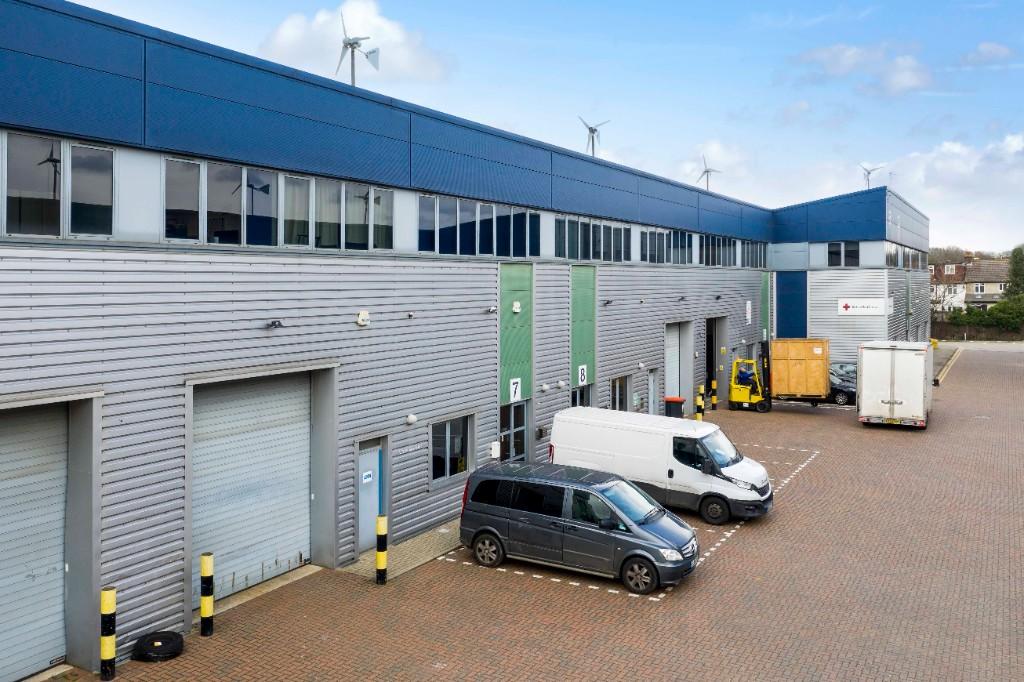 Industrial warehouse units with blue and grey exteriors, two parked delivery vans, a forklift, and a large crate on a paved lot under a partly cloudy sky.