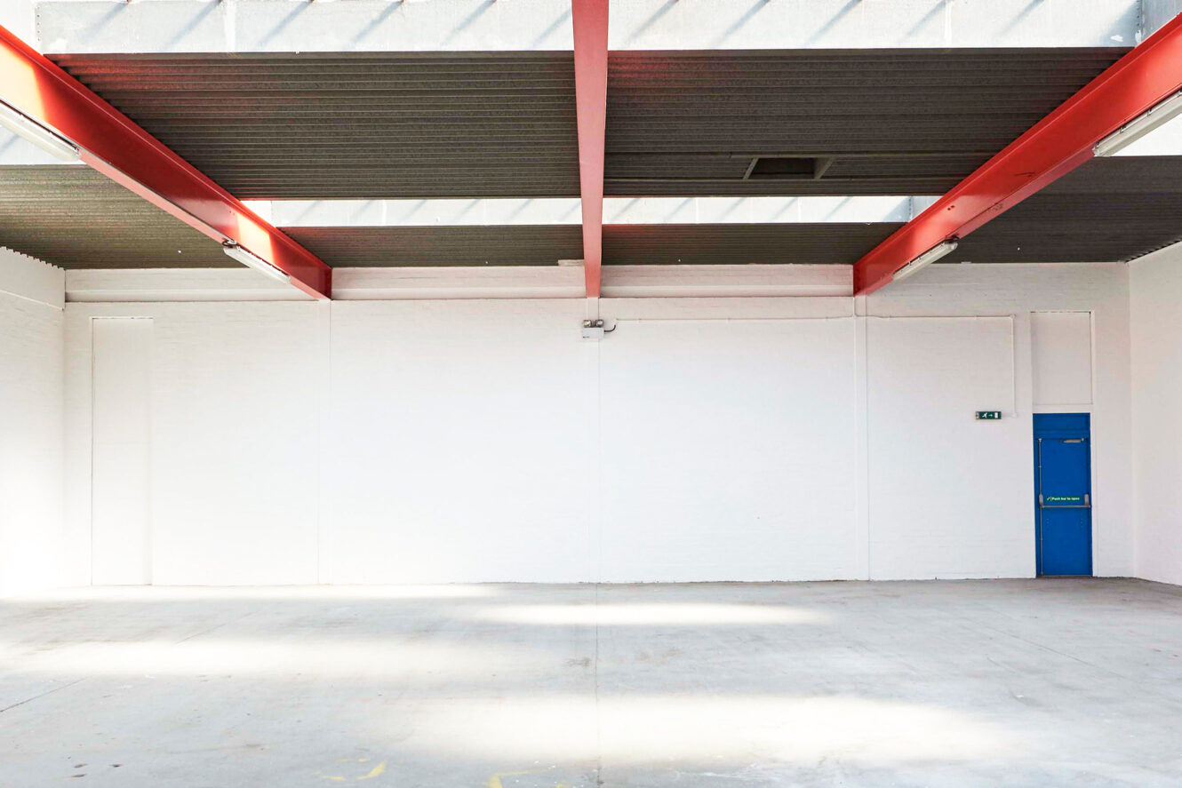 Empty industrial warehouse with red ceiling beams, white walls, concrete floor, skylights, and a blue exit door on the right side.