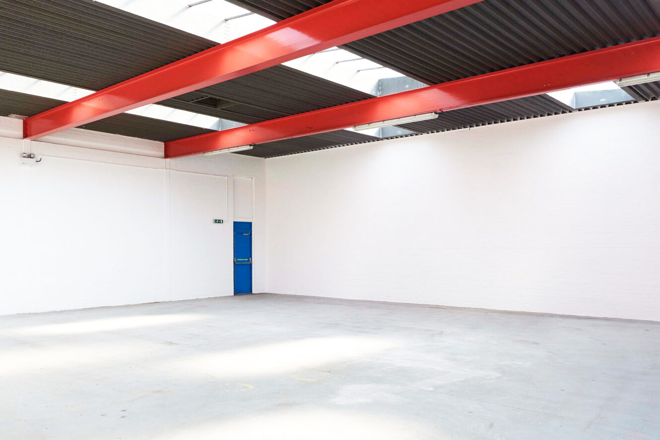 Empty warehouse interior with white walls, grey concrete floor, red ceiling beams, skylights, and a blue door in the corner.