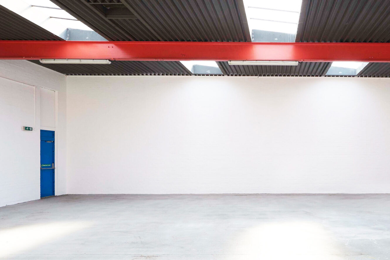 Empty industrial space with a concrete floor, white walls, a blue door, red ceiling beam, and skylights letting in natural light.