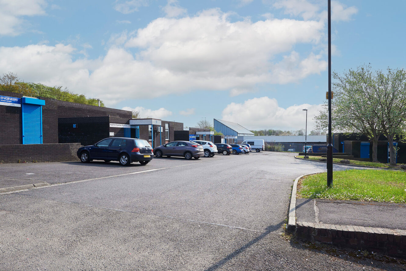 A parking lot with several parked cars beside low industrial buildings under a partly cloudy sky. A grassy area and trees are visible on the right side.