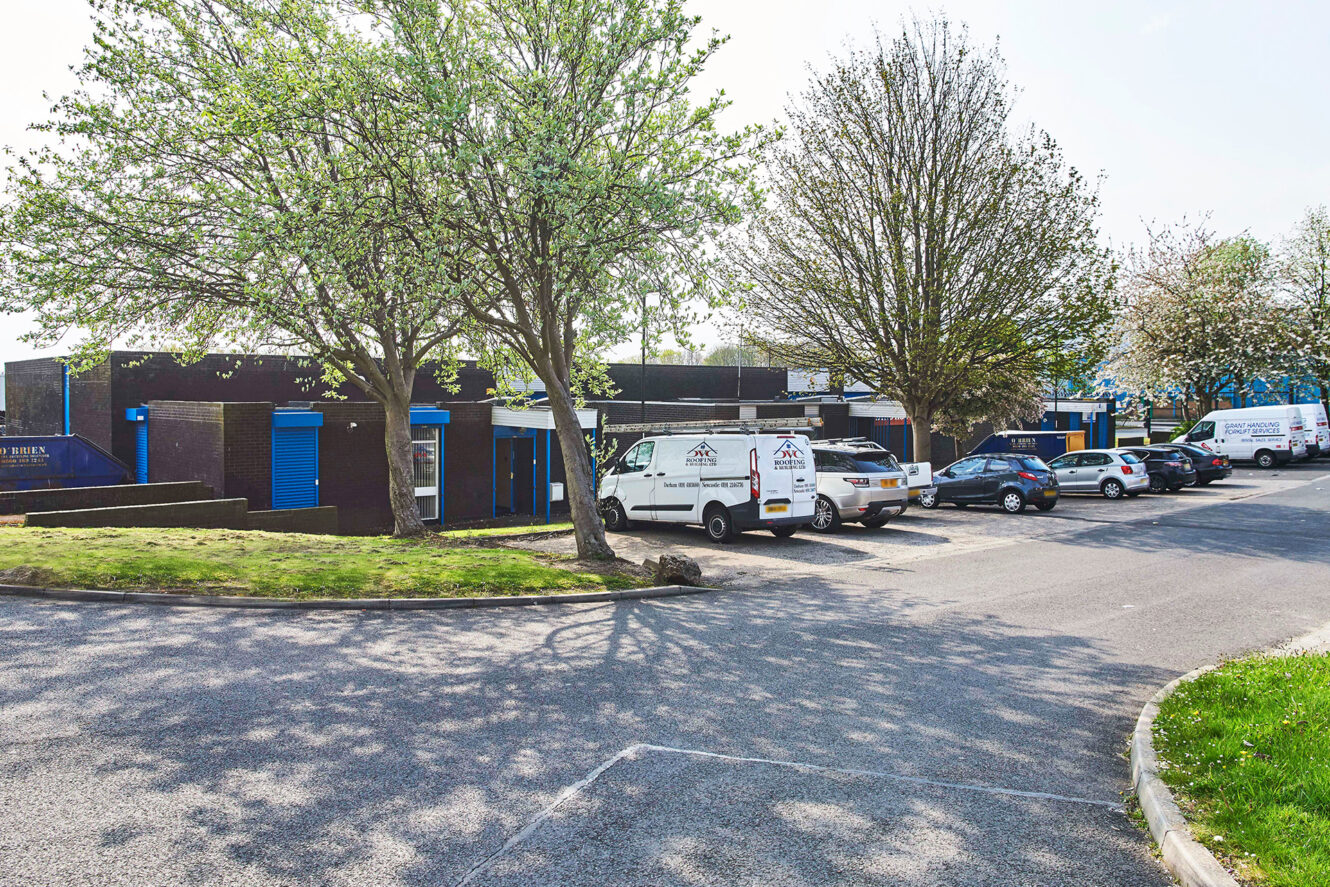 A small parking lot with cars and white vans outside a low, flat-roofed building surrounded by trees with green leaves.
