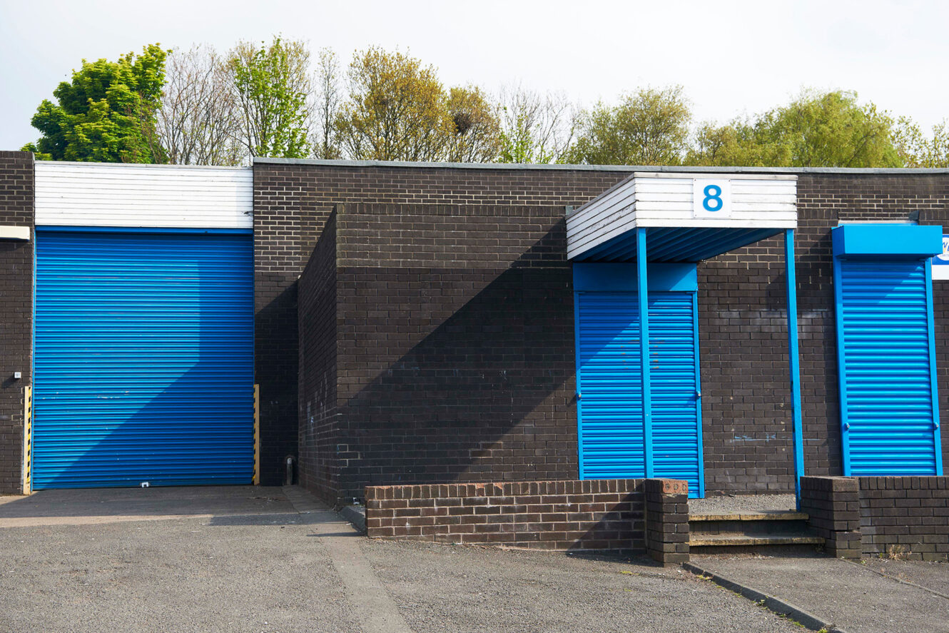 A row of brick industrial units with blue roller doors and canopies; one door is labeled with the number 8, and there are trees in the background.