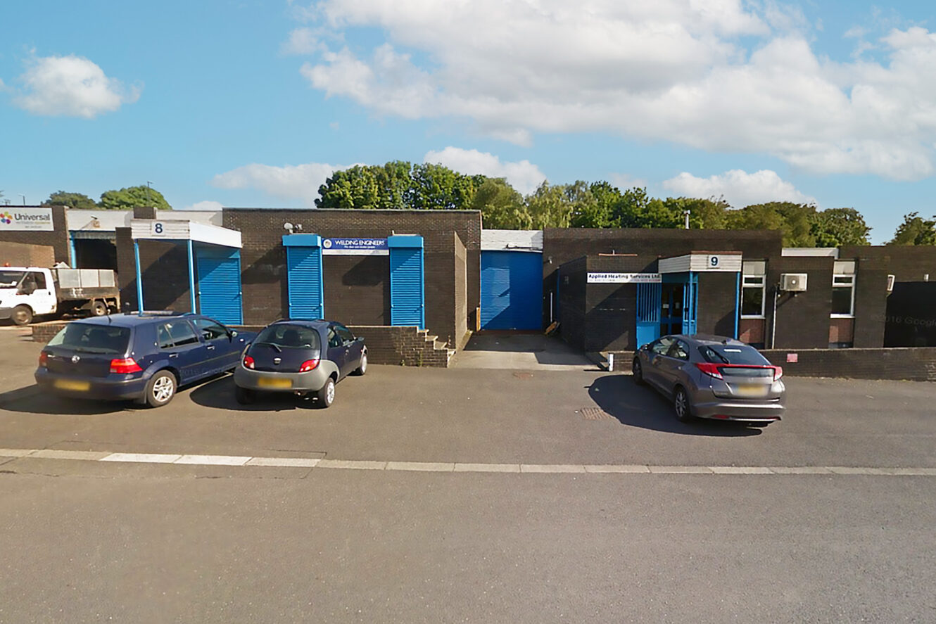 A row of industrial units with blue doors and numbers 8 and 9, three parked cars in front, and trees in the background under a partly cloudy sky.