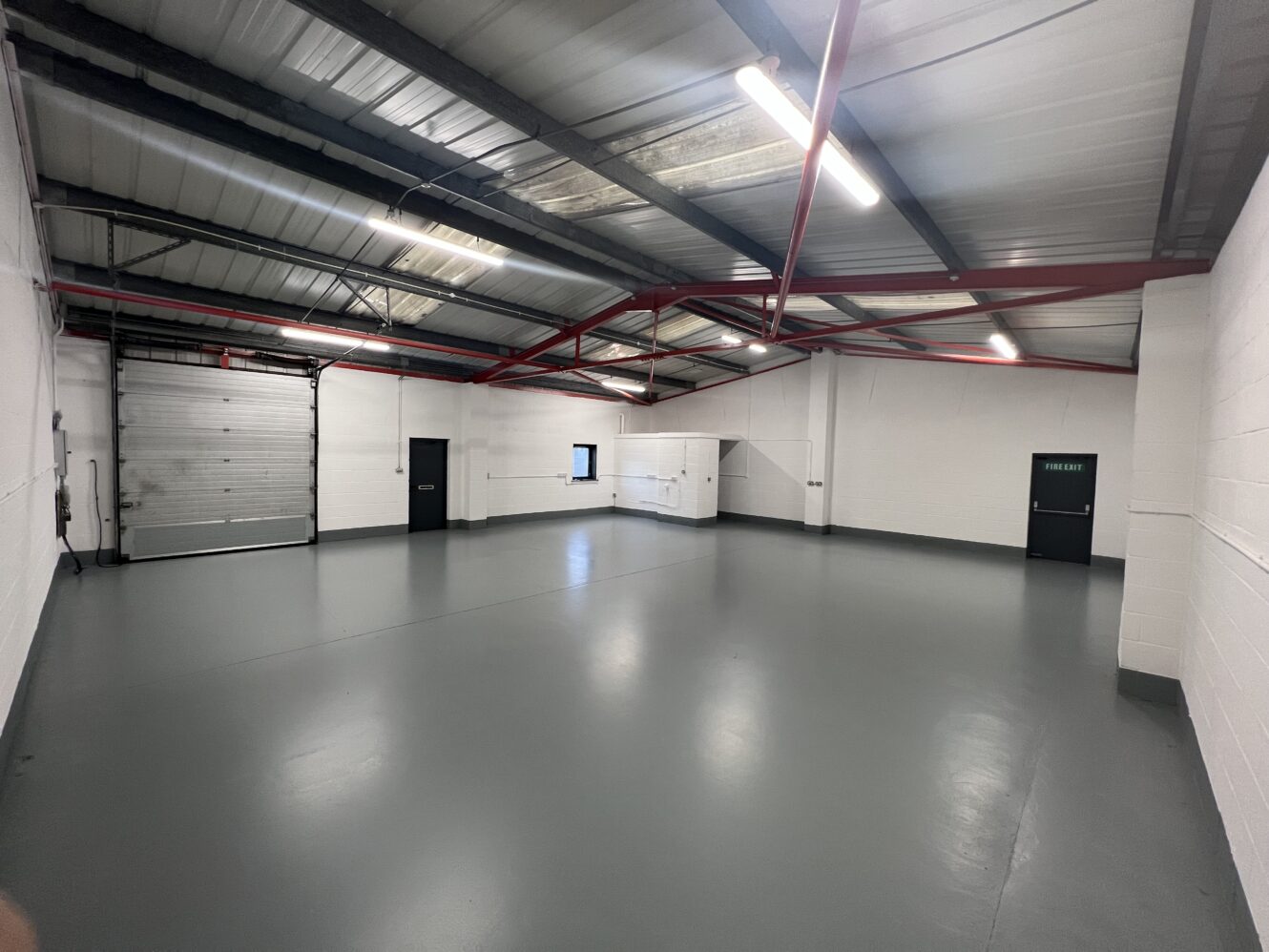 An empty industrial warehouse unit with gray concrete floor, white walls, exposed red steel beams, a large shutter door, and fluorescent ceiling lights.
