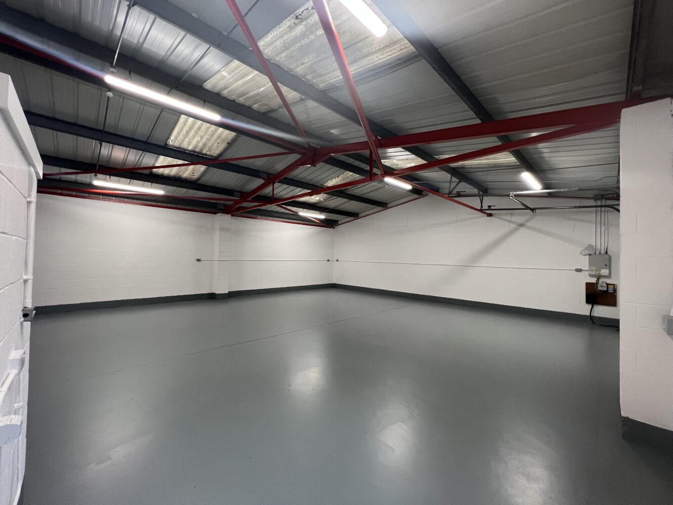 An empty industrial room with white walls, gray floor, and exposed red metal ceiling beams under fluorescent lighting.