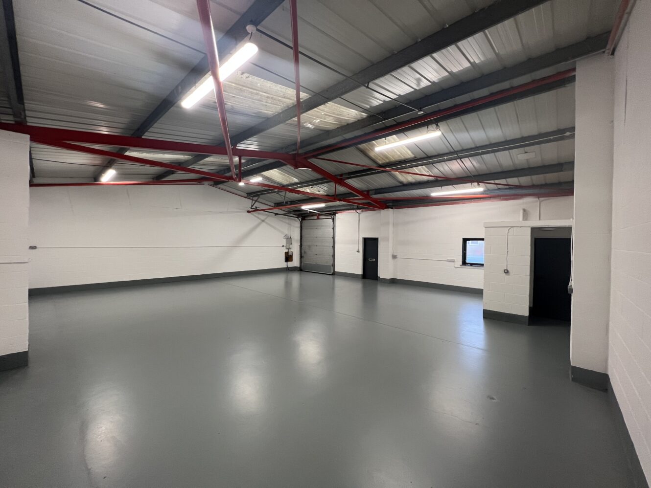 Empty industrial warehouse space with grey floor, white walls, exposed red ceiling beams, fluorescent lights, and a few doors and windows.