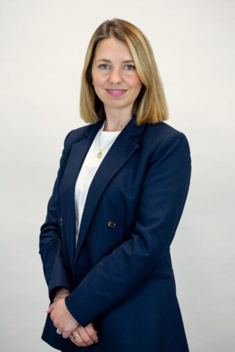 A woman with shoulder-length blonde hair wearing a navy blazer and white shirt stands against a plain light background, smiling slightly with her hands clasped in front.