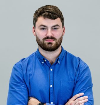 A man with short brown hair and a beard, wearing a blue button-up shirt, stands with arms crossed against a plain grey background.