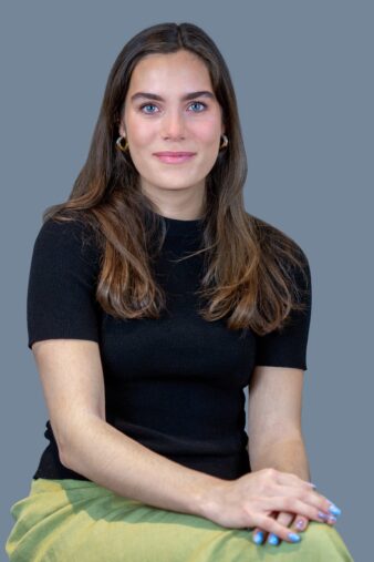 A woman with long brown hair, wearing a black short-sleeve top and light green skirt, sits with her hands folded on her lap against a plain gray background.