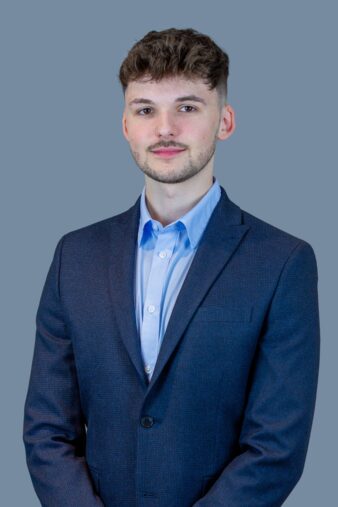 A young man with short brown hair and a trimmed beard, wearing a blue suit jacket and light blue shirt, stands against a plain gray background.