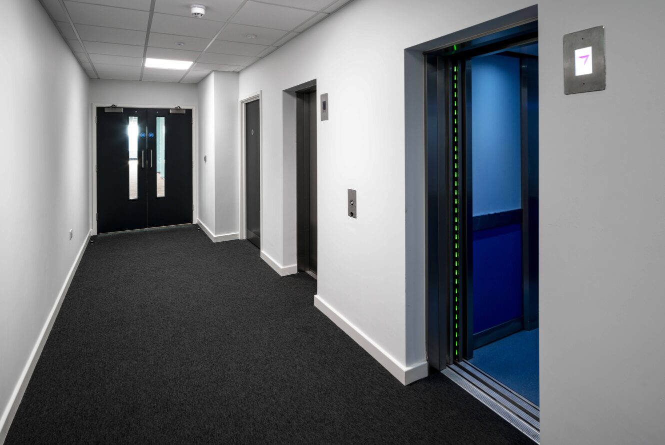 A modern hallway with two elevators; one elevator door is open with blue light inside, and double doors are visible at the end of the carpeted corridor.