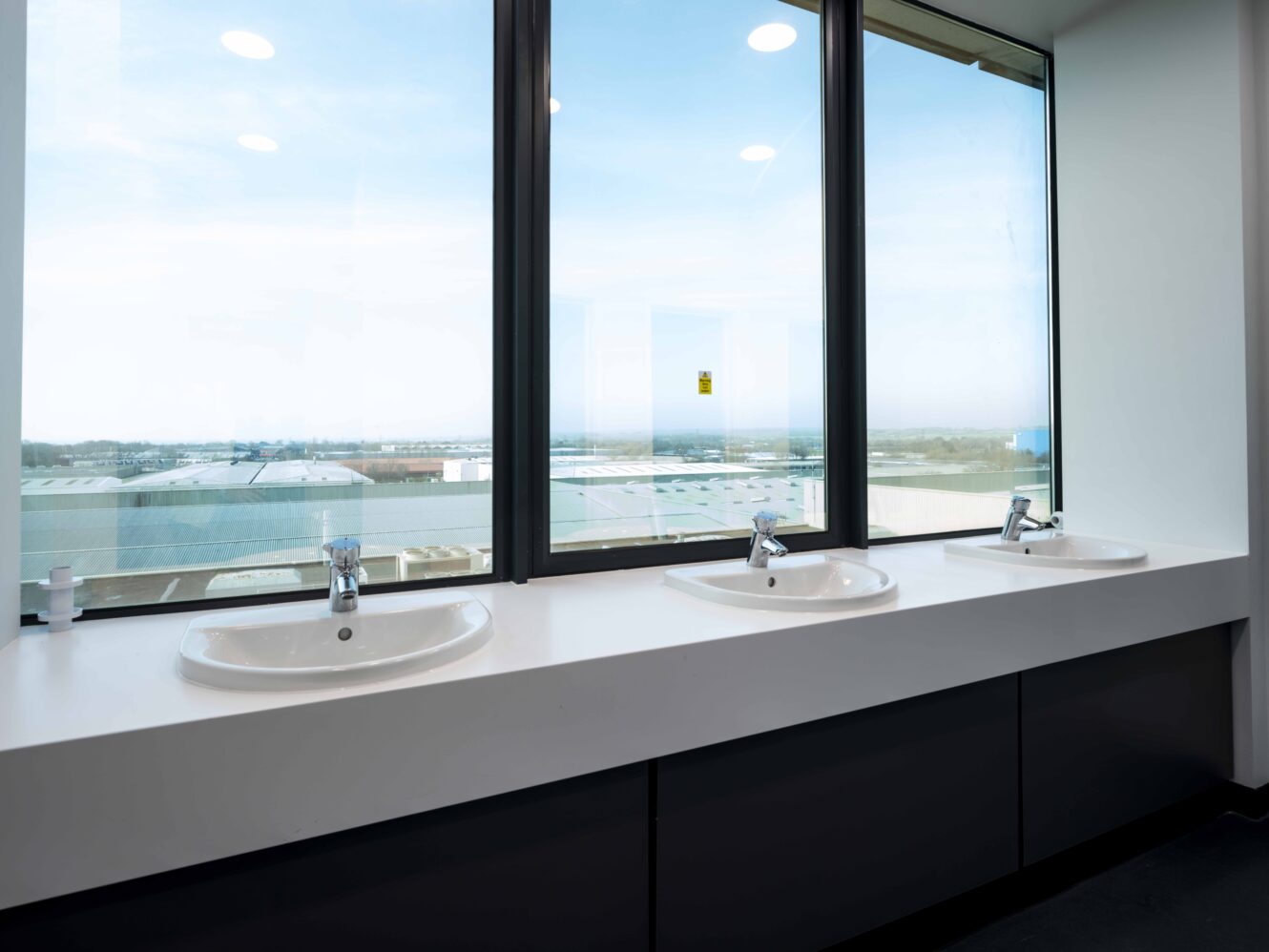 Three white sinks with chrome faucets are installed on a countertop in front of large windows overlooking an industrial area and a blue sky.