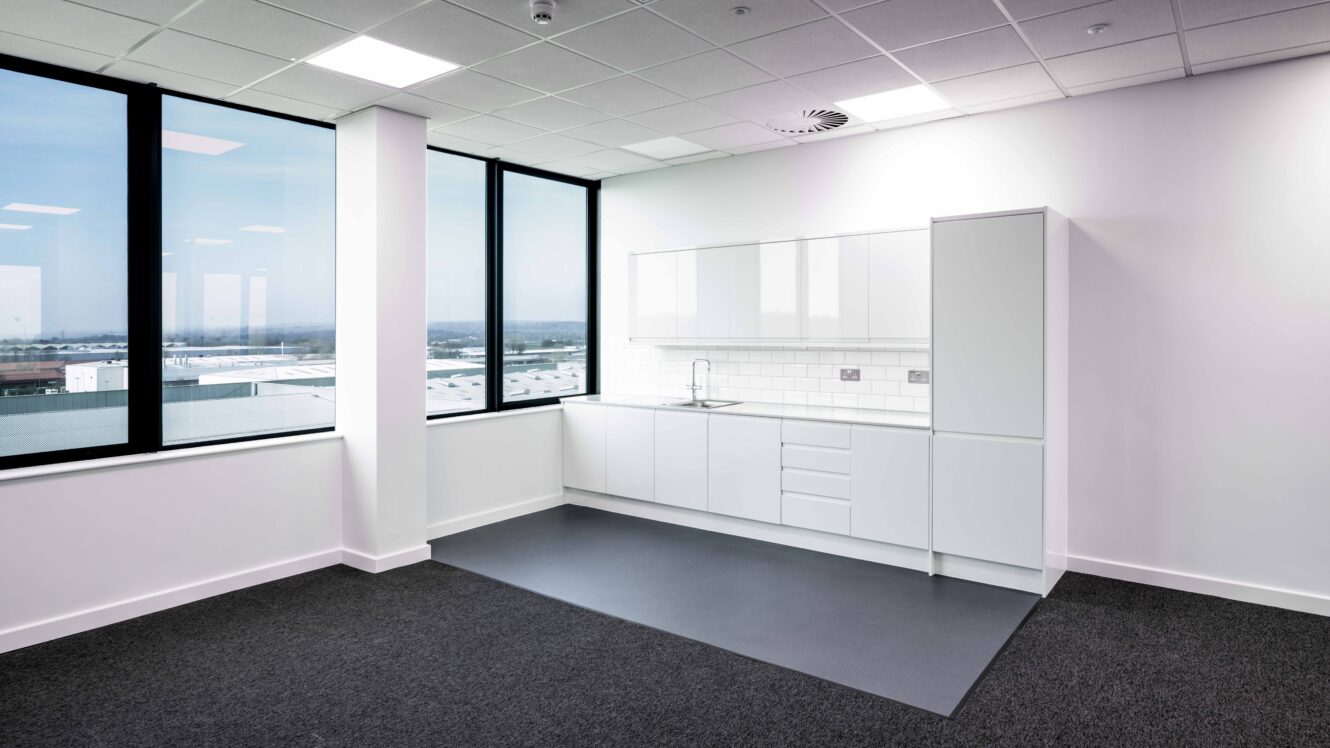 Modern office kitchenette with white cabinets, sink, and tile backsplash, situated in a corner with large windows and black carpeted floor.
