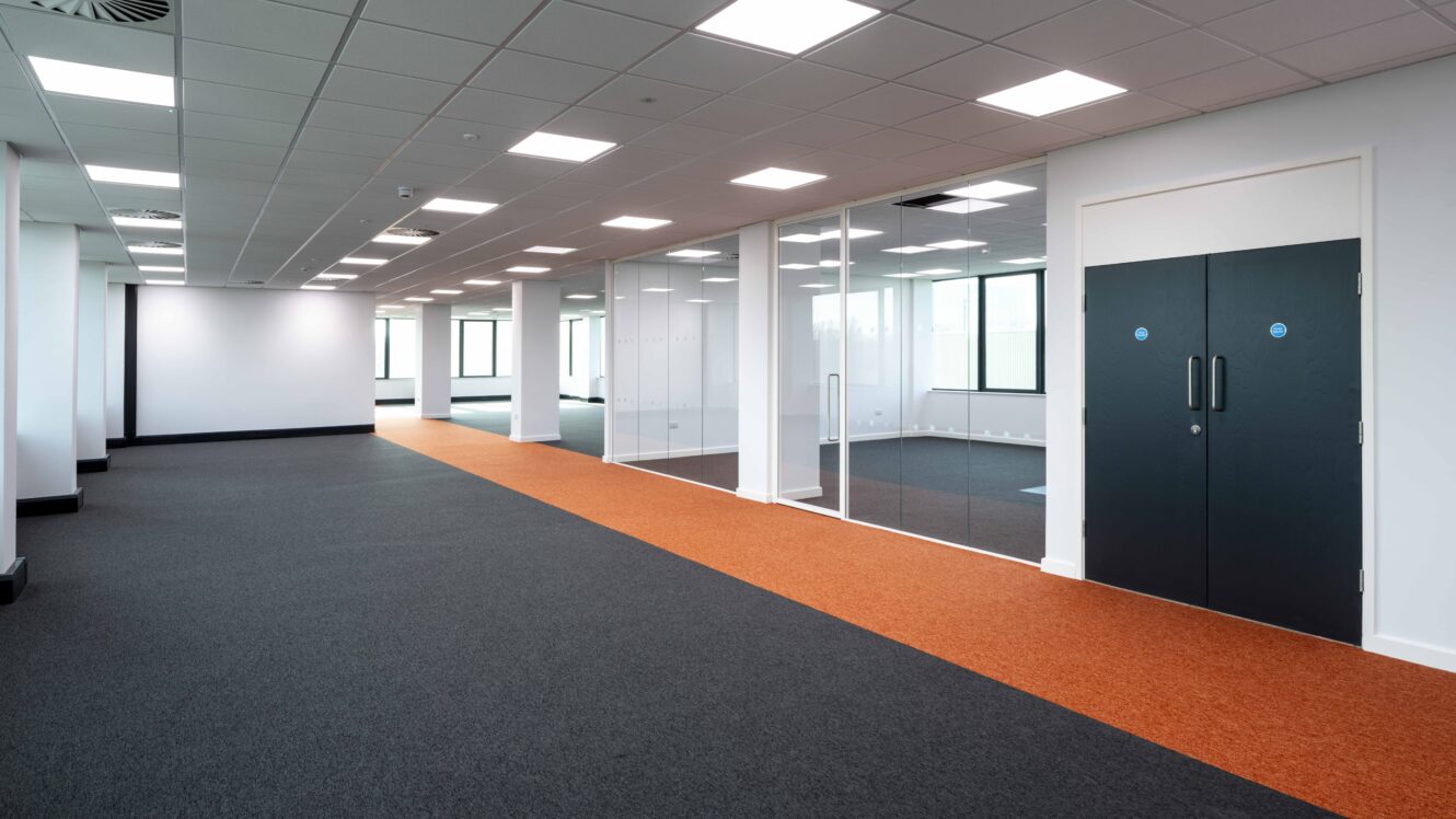 Modern empty office space with a glass-walled meeting room, black double doors, white walls, ceiling lights, and a bright orange carpeted walkway.