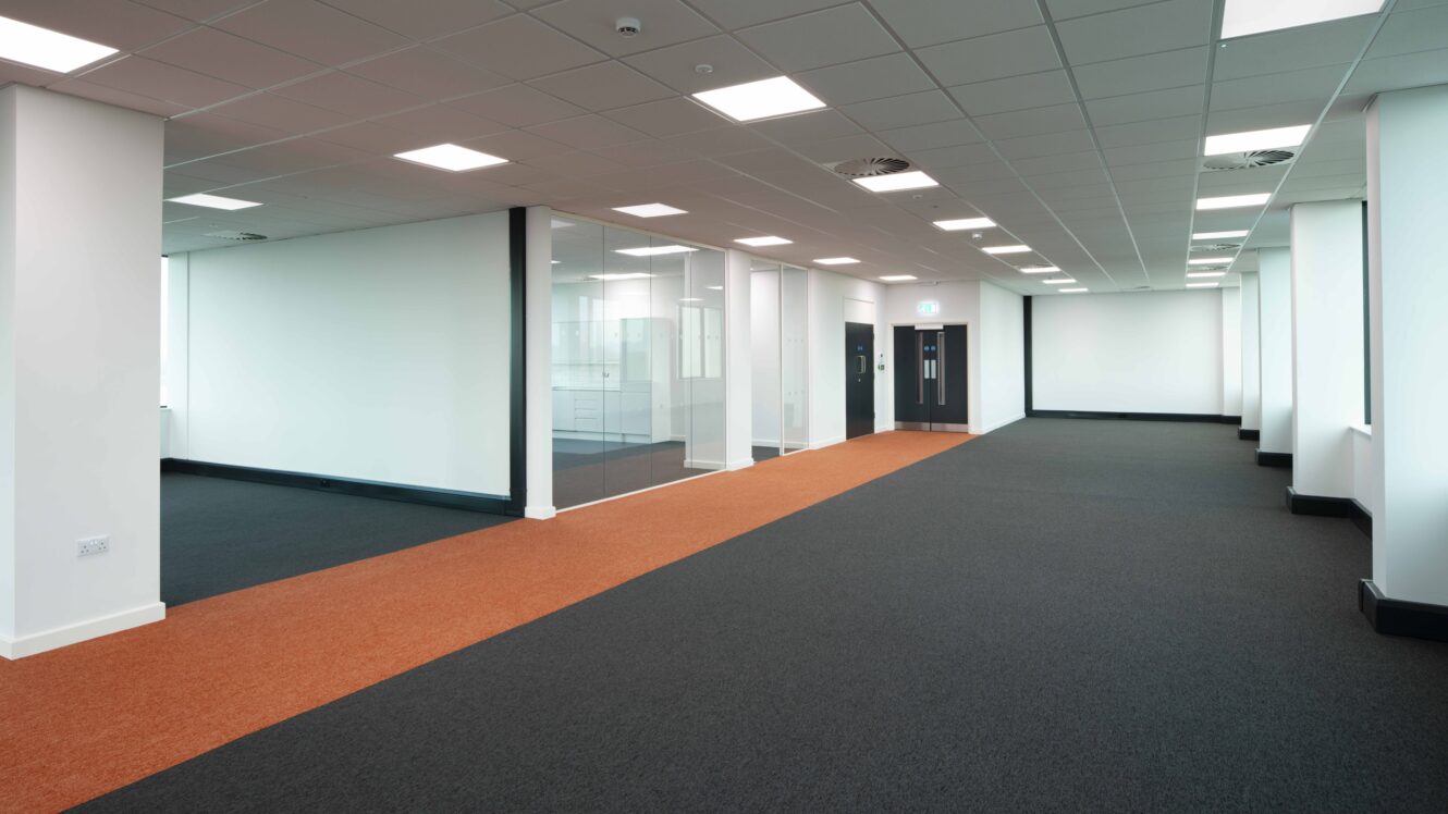 Modern empty office space with a glass-walled room, white walls, ceiling lights, and carpeting in black and orange sections.