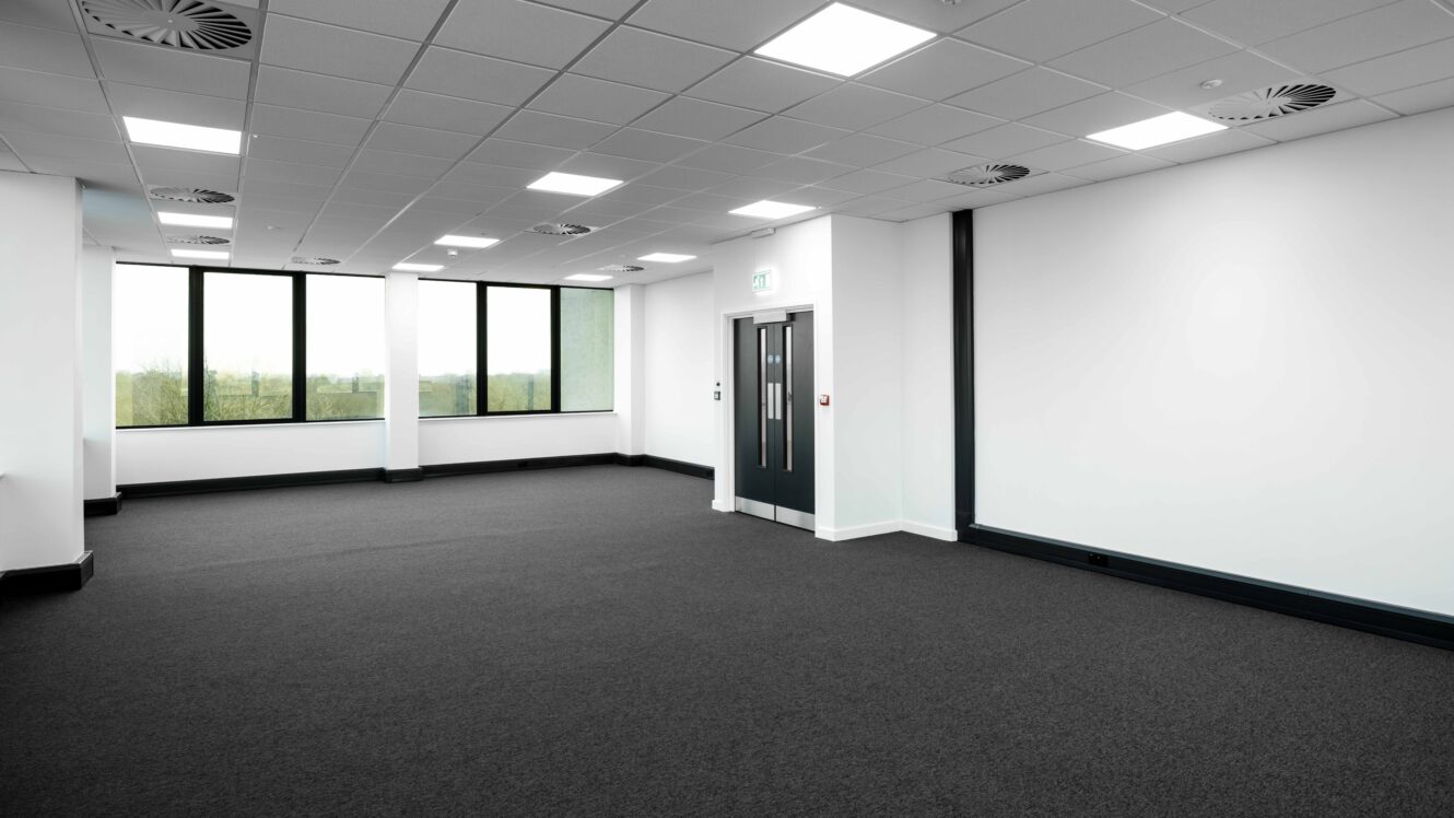 Empty office space with white walls, grey carpet, large windows, and double doors under fluorescent ceiling lights.