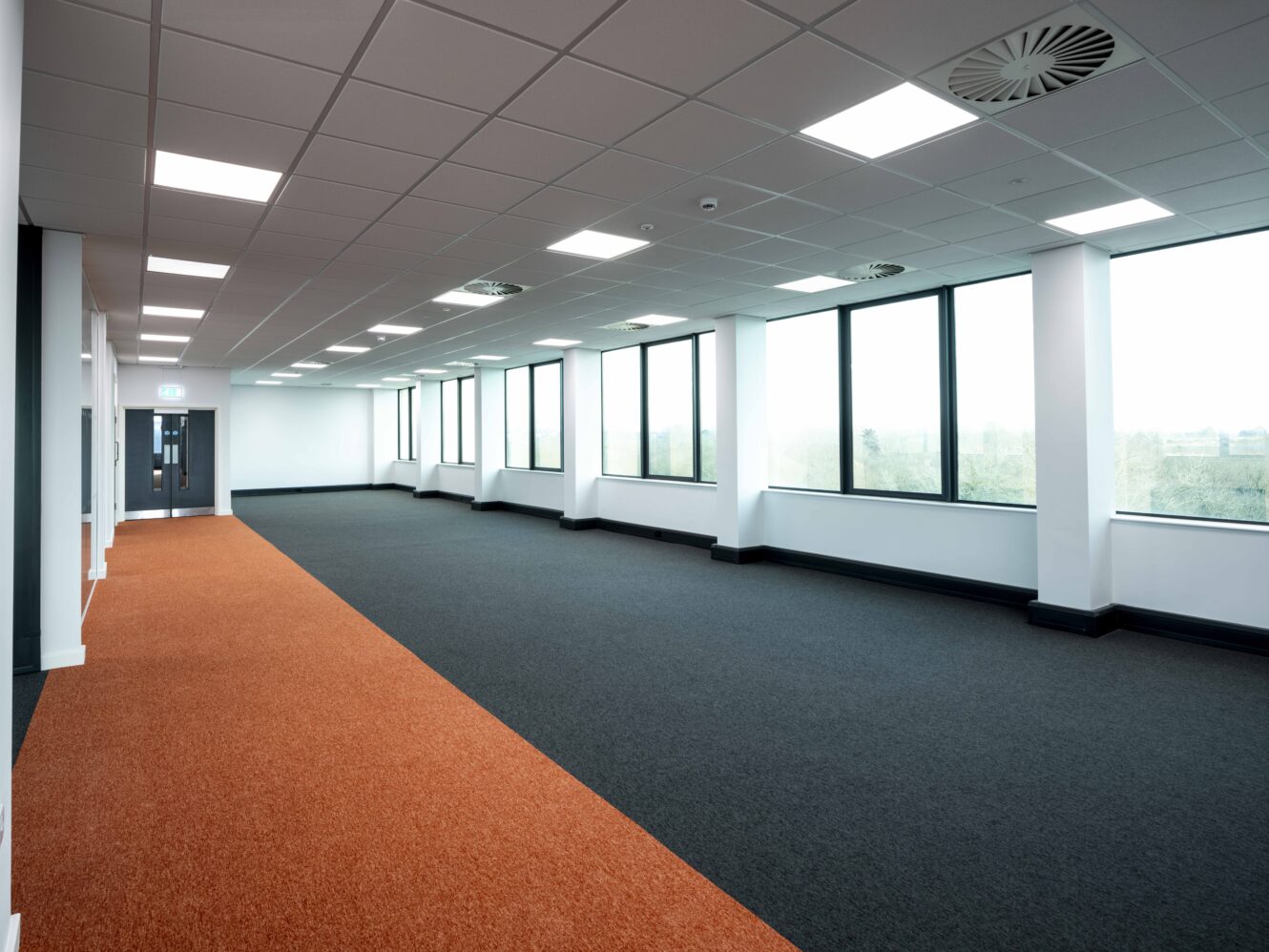 Empty modern office space with large windows, white walls, and a ceiling with fluorescent lights. The floor is partly covered in orange and black carpet tiles.