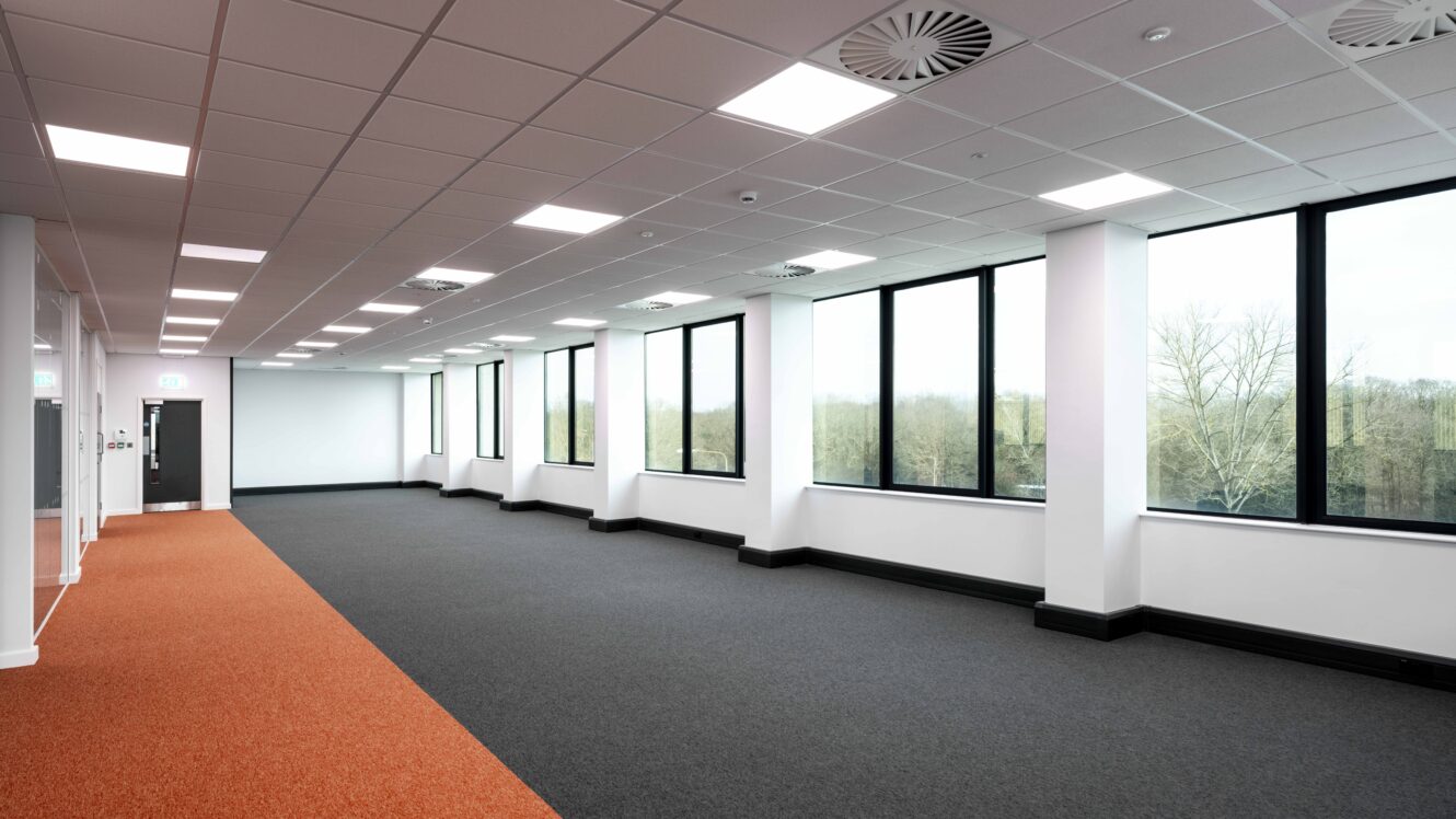 Empty modern office space with large windows, white walls, a black and orange carpet, and ceiling lights. Trees are visible outside through the windows.