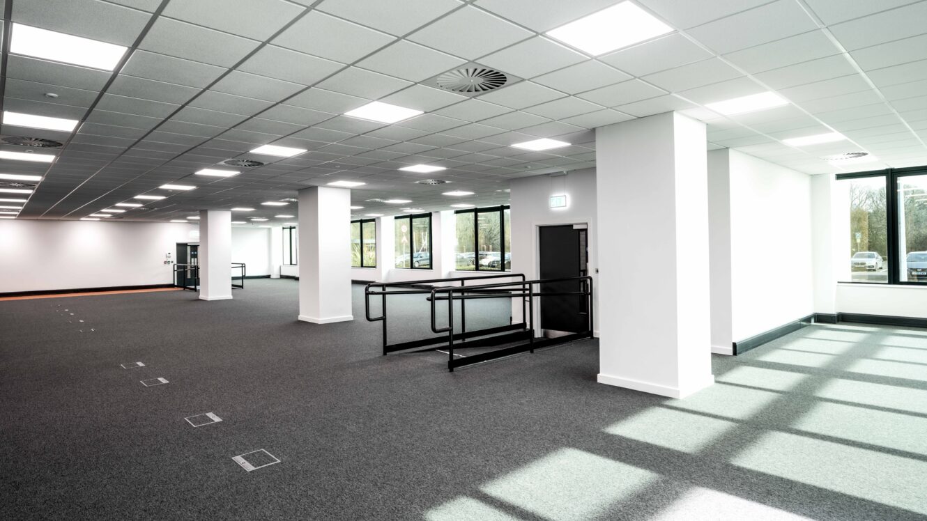 A modern, empty office space with large windows, white walls, carpeted floor, and overhead fluorescent lighting. There is a black accessible ramp near the center.