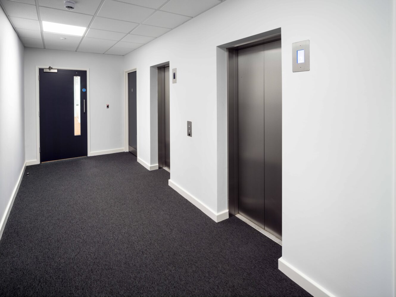 A hallway with two closed elevator doors on the right, a closed door at the end, and white walls with a gray carpeted floor.