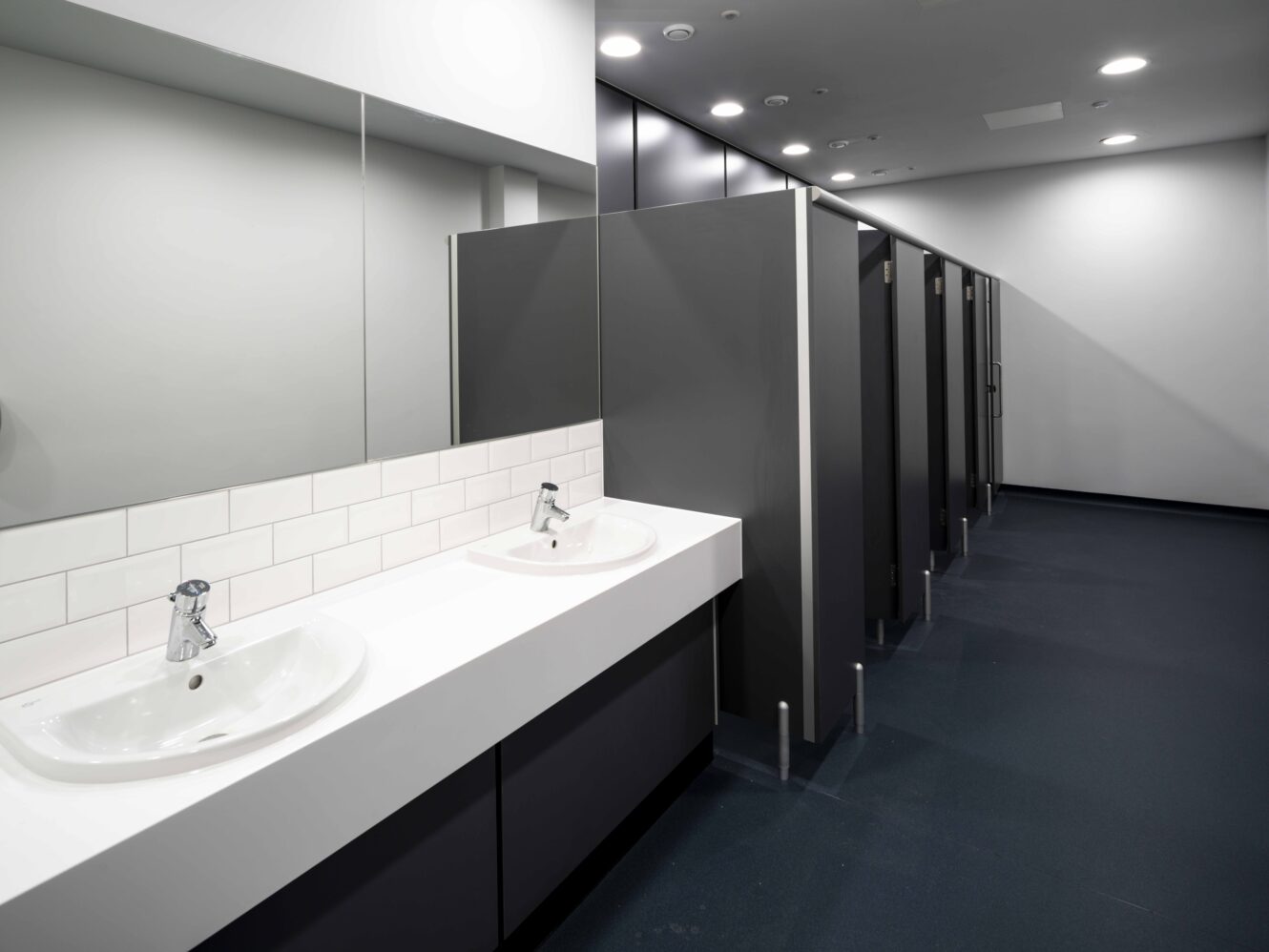 Modern public restroom with two white sinks, a large mirror above, and a row of closed dark gray toilet stalls on the right.