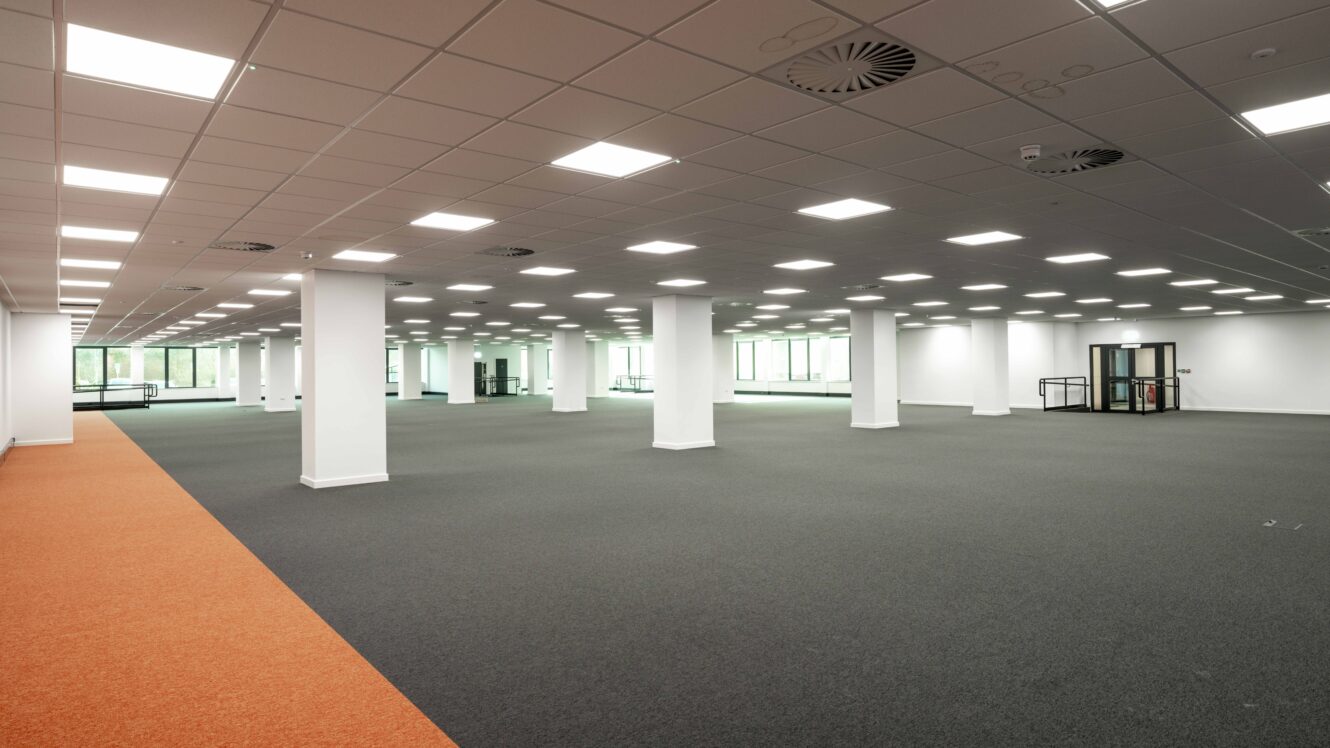 Large, empty office space with gray carpet, white columns, orange walkway, ceiling lights, and windows along the far walls.