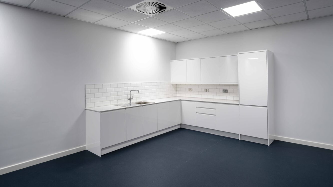 Minimalist kitchen with white cabinets, tiled backsplash, stainless steel sink, dark floor, and overhead fluorescent lighting.