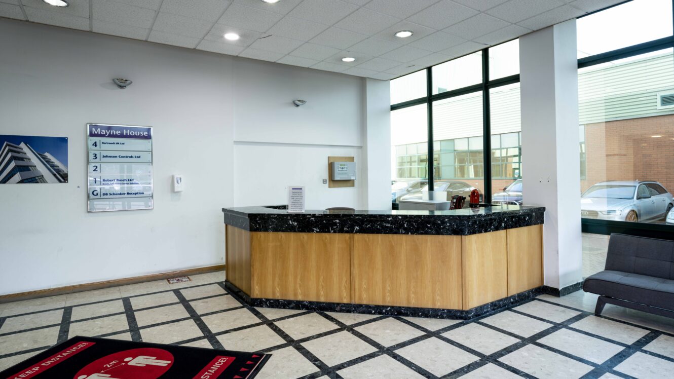 A modern building reception area with a wooden front desk, tiled floor, large windows, and a wall directory sign labeled Mayne House.