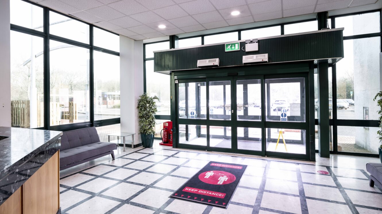 Modern building lobby with large glass doors, a gray sofa, marble floor, potted plants, and a red floor sign reminding visitors to keep distance.