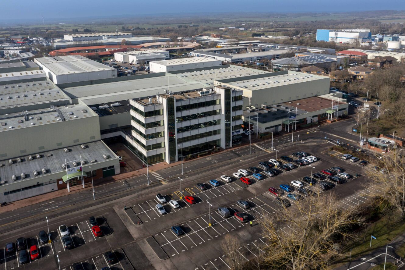 Aerial view of a large industrial complex with office buildings, warehouses, and a parking lot filled with cars. Trees line the edge of the lot, and more industrial buildings are visible in the background.