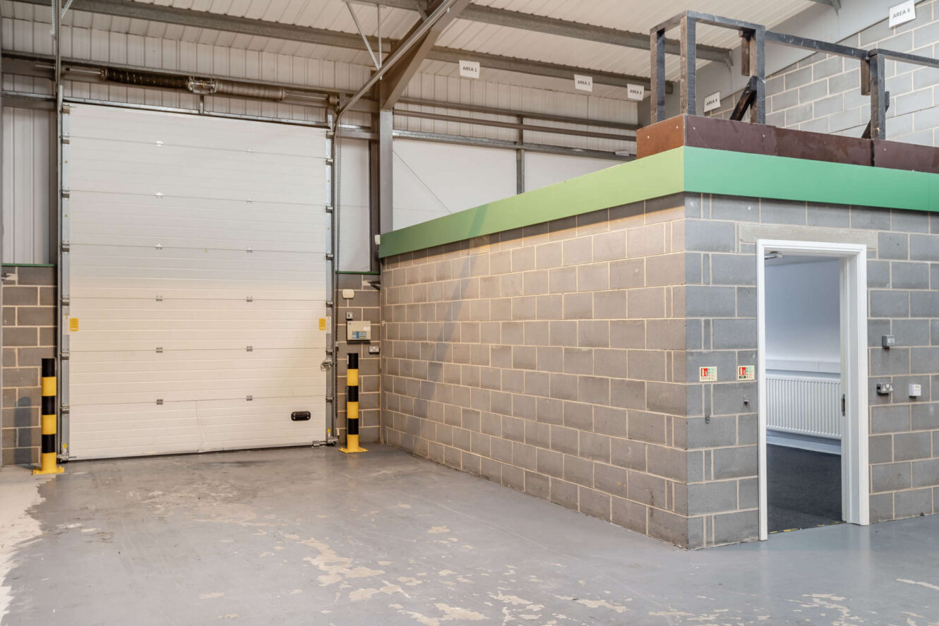 Industrial warehouse interior with exposed concrete block walls, a large shutter door, a small mezzanine, and an office entrance with an open door.