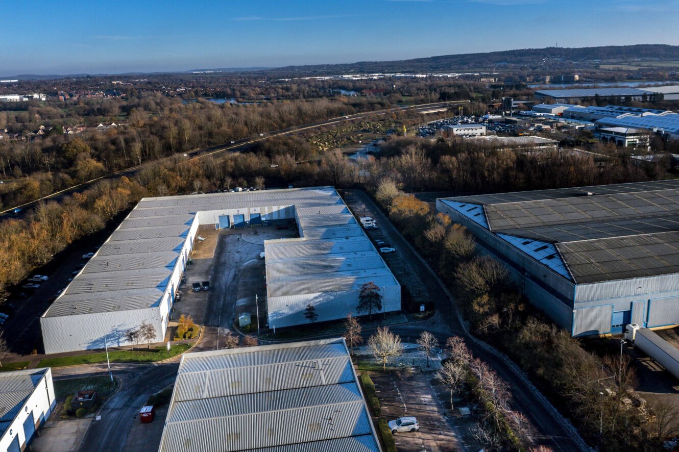 Aerial view of several large industrial warehouse buildings surrounded by trees, roads, and parking areas on a clear day.
