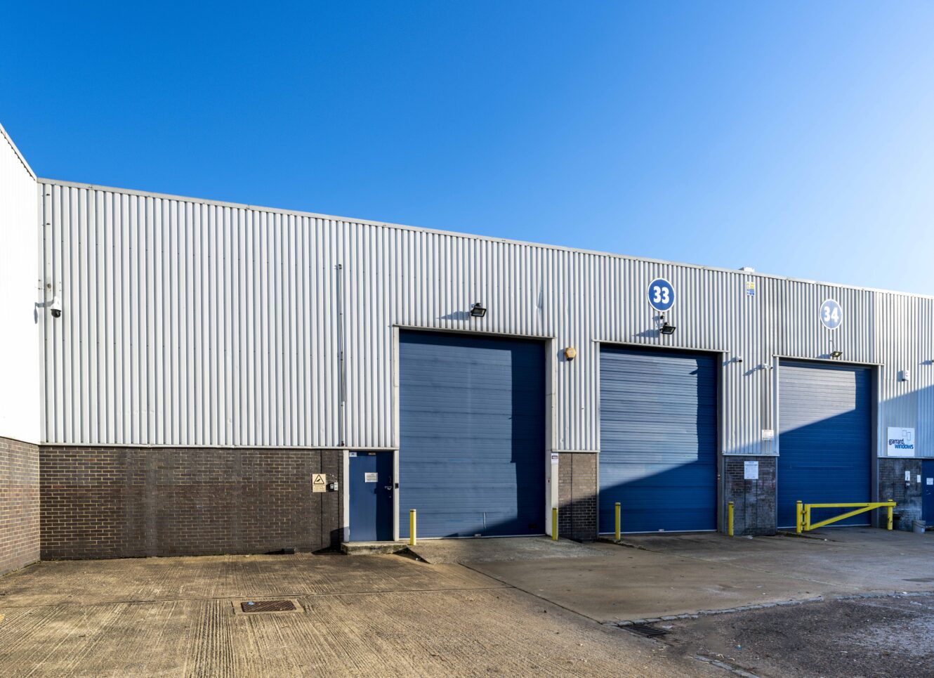 Industrial warehouse exterior with three large blue roller shutter doors numbered 33 and 34, under a clear blue sky. Concrete yard in foreground.