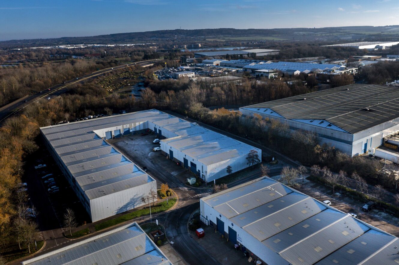 Aerial view of an industrial park with several large warehouse buildings, parking areas, and surrounding trees under a clear blue sky.