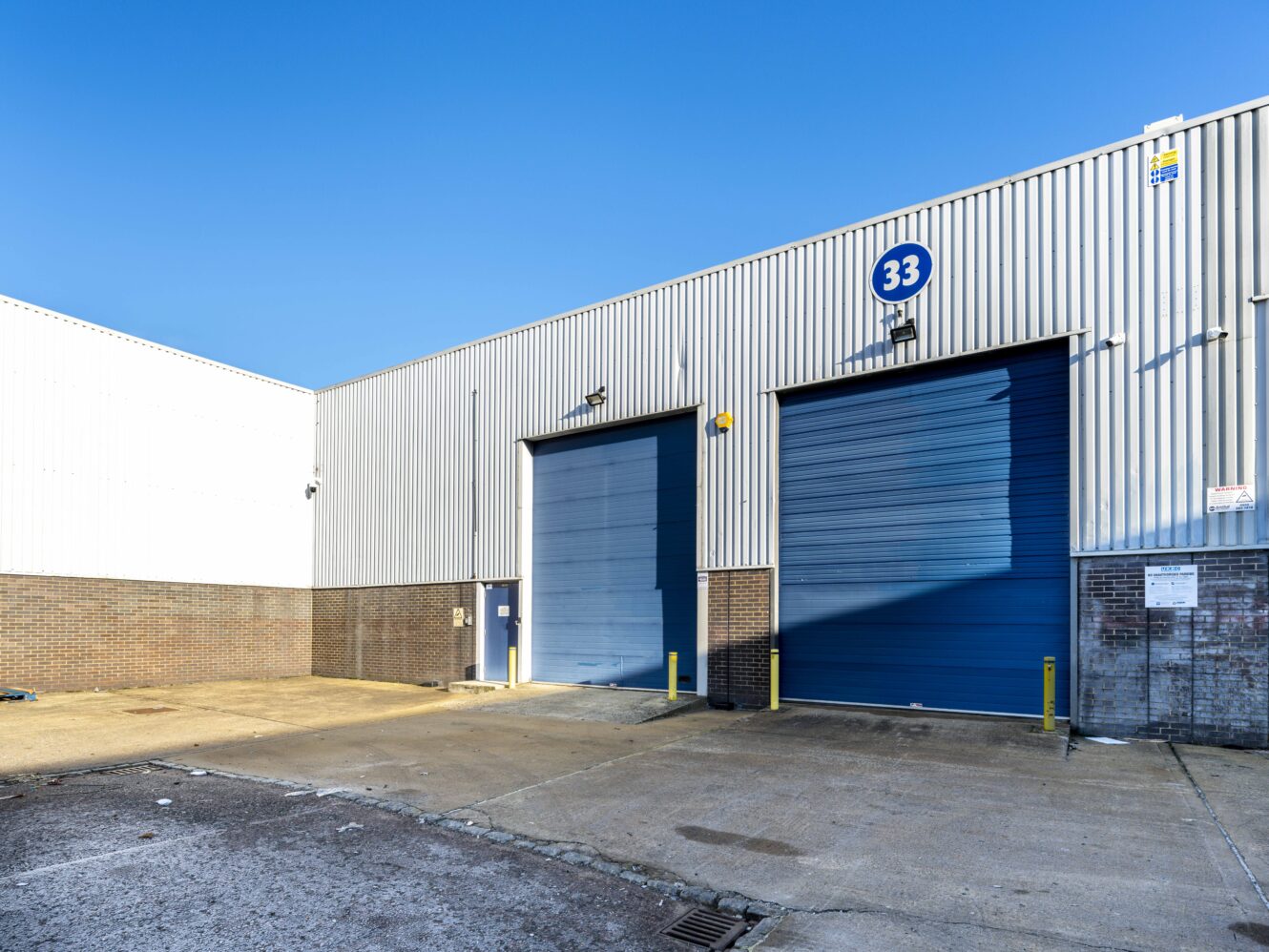 Industrial warehouse with two large blue roller doors, marked as unit 33, under a clear blue sky. Concrete yard in foreground.