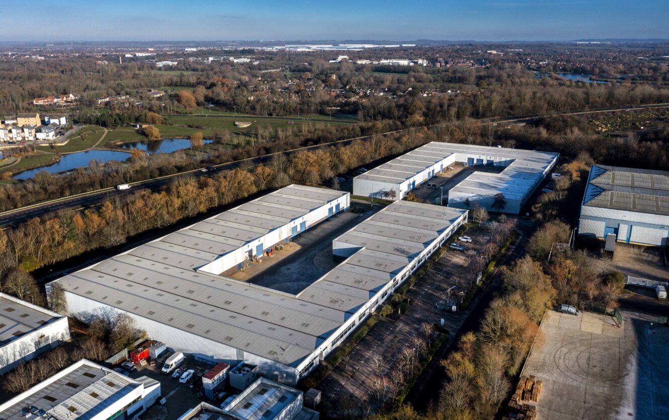 Aerial view of a large industrial complex with multiple white-roofed warehouse buildings, surrounded by trees, roads, and open land.