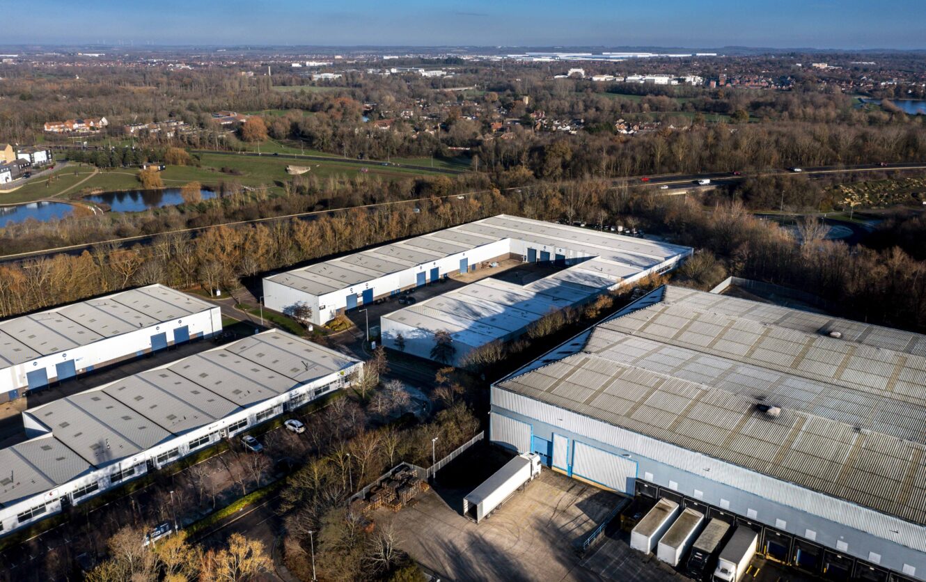 Aerial view of a large industrial warehouse complex with multiple buildings, loading docks, parked trucks, and surrounding trees near a suburban area.