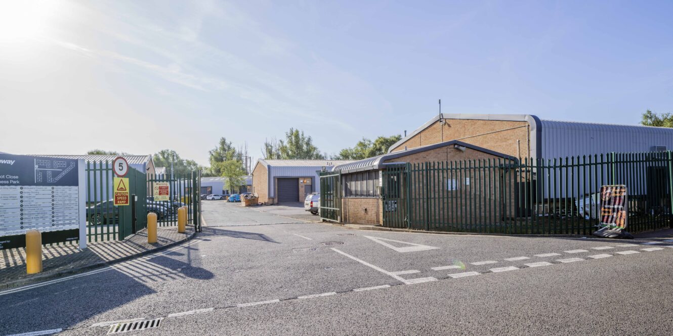 Industrial facility with fenced perimeter, security gate, warning signs, and several low brick and metal buildings visible inside under a clear sky.