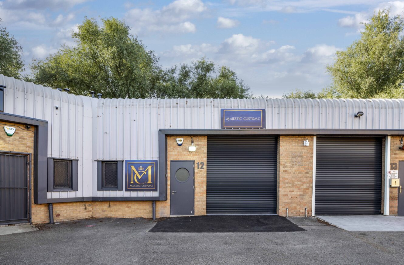 A small industrial building with two black roller doors, a grey door numbered 12, and signs reading Majestic Customs on the front. Trees and blue sky in the background.