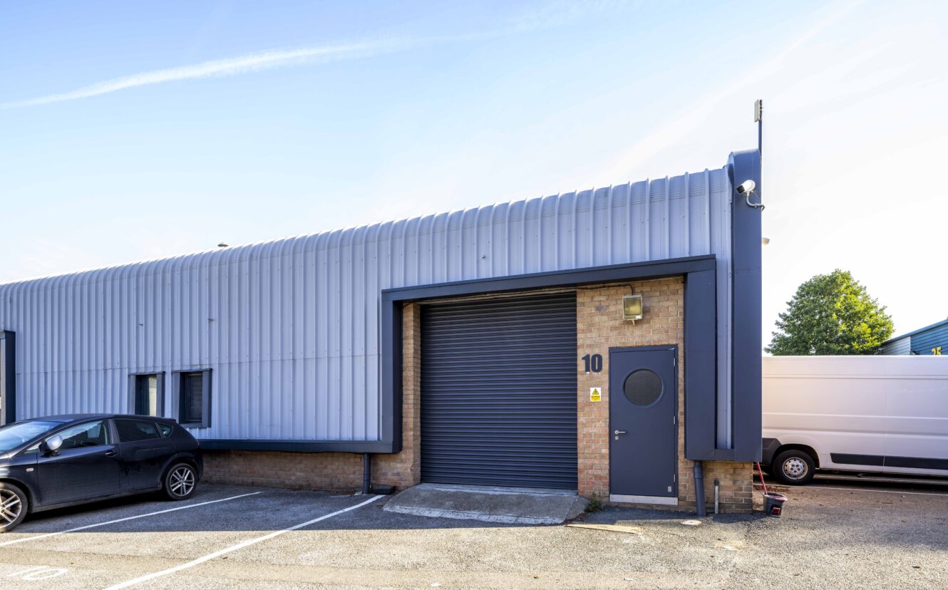 A small industrial warehouse unit with a blue exterior, a large roller shutter door, a standard door marked 10, and cars parked outside.