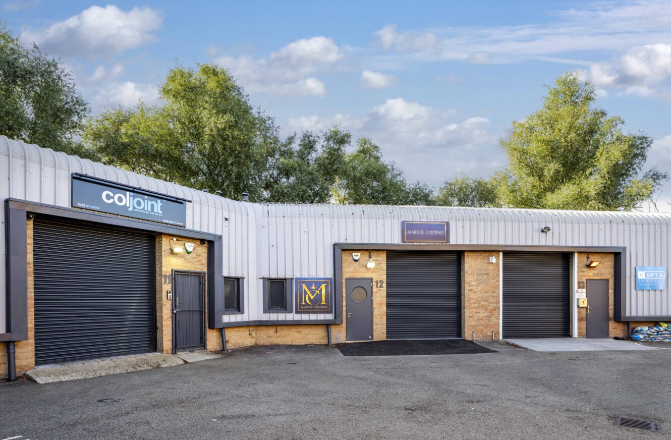 Single-story industrial building with three roller shutter doors, brick and metal exterior, and three business signs, set against a backdrop of trees and partly cloudy sky.