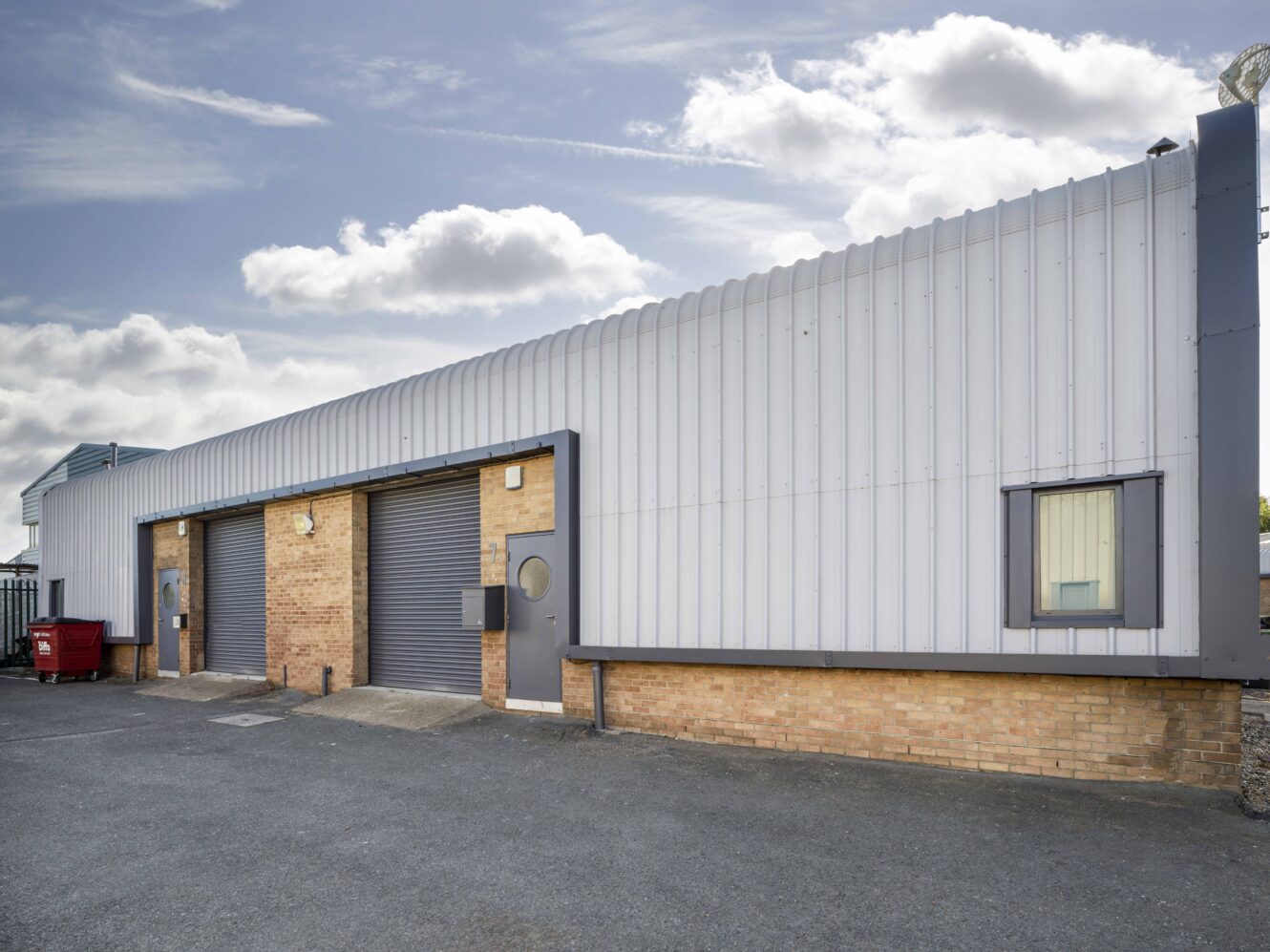 A single-story industrial warehouse with a metal roof, brick lower walls, three roller shutter doors, and one window, set on a paved lot under a partly cloudy sky.